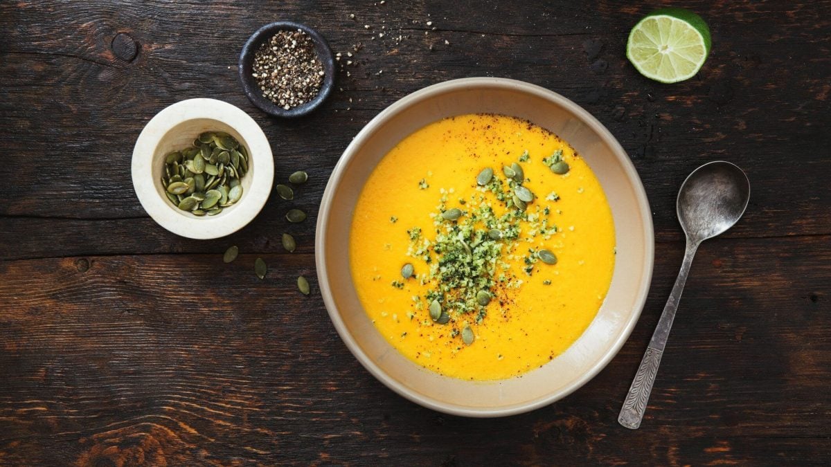 Kalte Gemüsesuppe mit Gurken, Möhren und Paprika, bestreut mit Körnern und Samen, auf dunklem Untergrund in der Draufsicht, daneben stehen einige Zutaten.