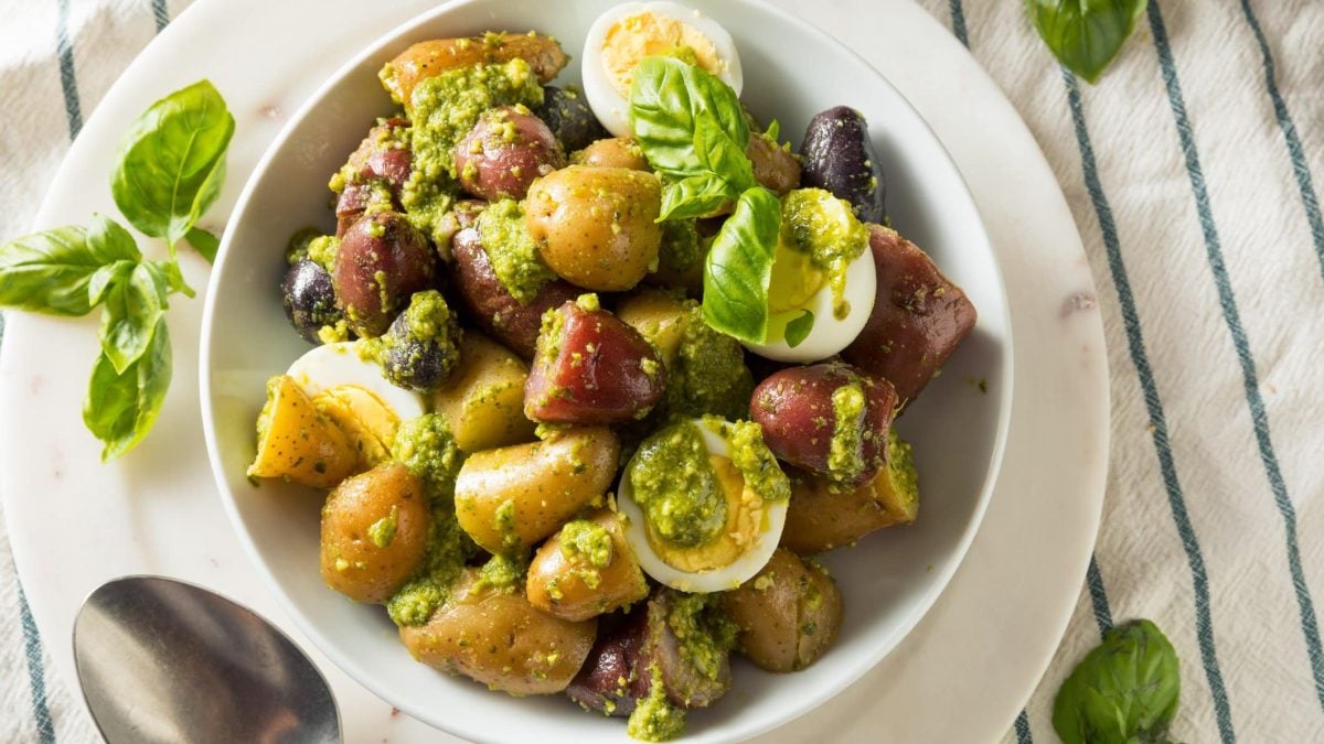Kartoffelsalat mit Pesto, Kalamata Oliven und Ei in weißer Schale auf weißen Teller auf blau-weißem Tischtuch. Basilikumblätter als Deko auf dem Salat und daneben im Bild. Metalllöffel unten links erkennbar. Aufnahme aus der Vogelperspektive.