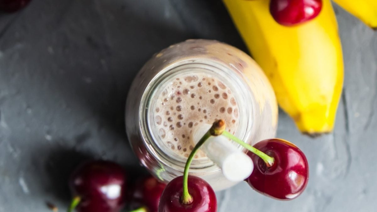 Ein Milkshake von oben fotografiert mit Kirschen und Bananen drum herum auf einem grauen Untergrund.