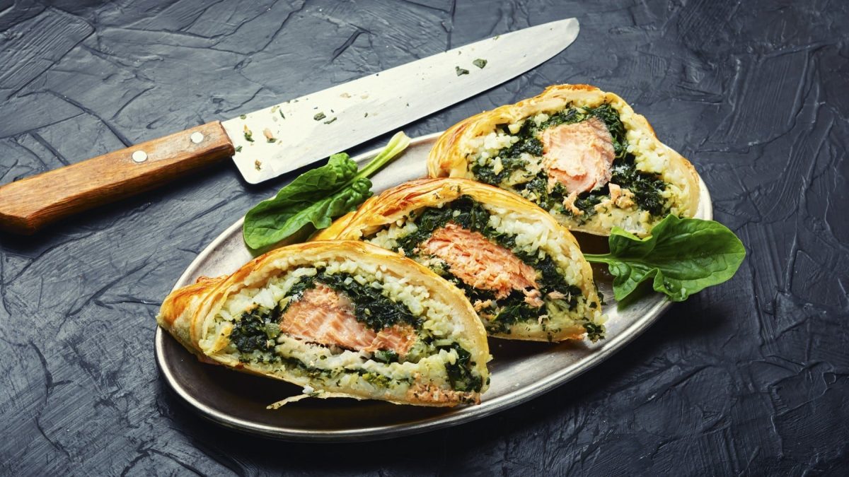 Spinat, Reis und Lachs im Teigmantel, in einzelne Portionen geschnitten auf einem hellen Teller, auf einem steinernen Tisch.