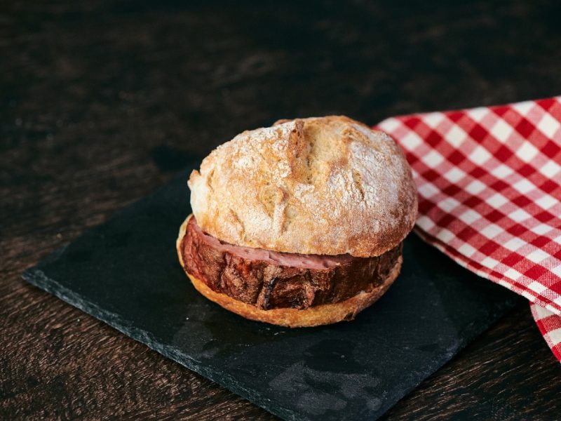 Brötchen mit Leberkäse innen auf einer Schieferplatte neben einem rot karierten Tuch