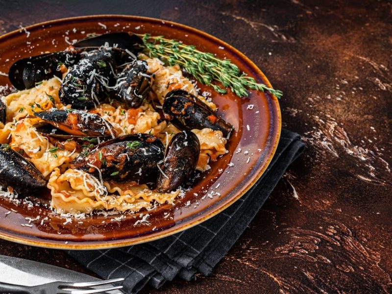 Mafaldine mit Miesmuscheln an einer Tomatensauce auf einem braunen- Teller und einem dunkelbraunem Holztisch.