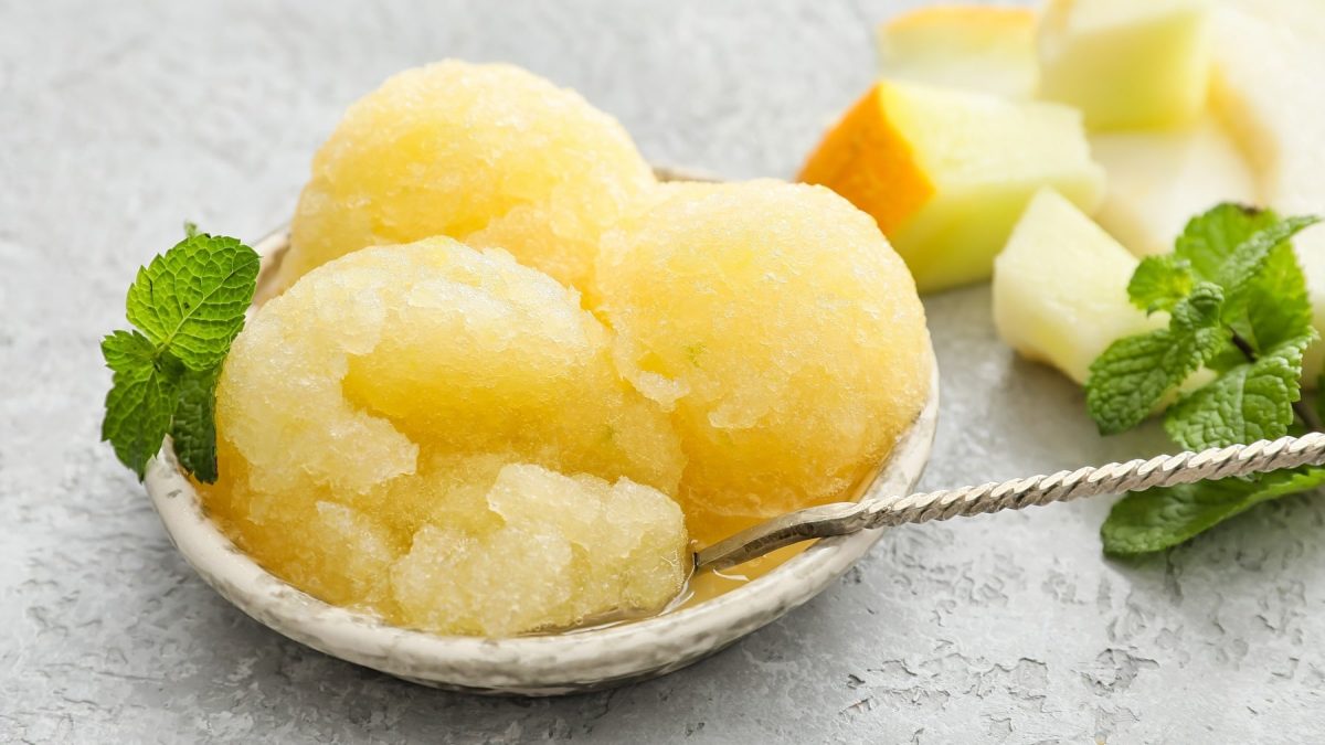 Ein Schälchen Melonensorbet mit Gin und einem Löffel, daneben liegen Minze und Melonenstücke.