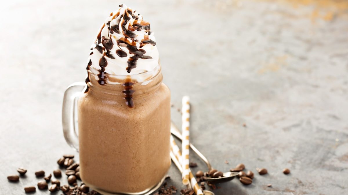 Ein Glas Mocha Frappuccino mit Schlagsahne und Schokosauce, daneben liegen Kaffeebohnen, Löffel und Strohhalme.