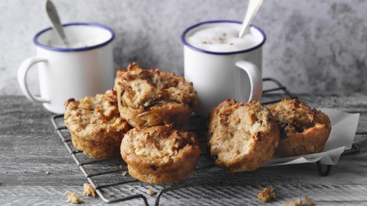 Fünf gebackene Müsli-Muffins auf einem Gitter und zwei helle Kaffeetassen im Hintergrund. Alles auf einem Holztisch und vor grauer Wand.