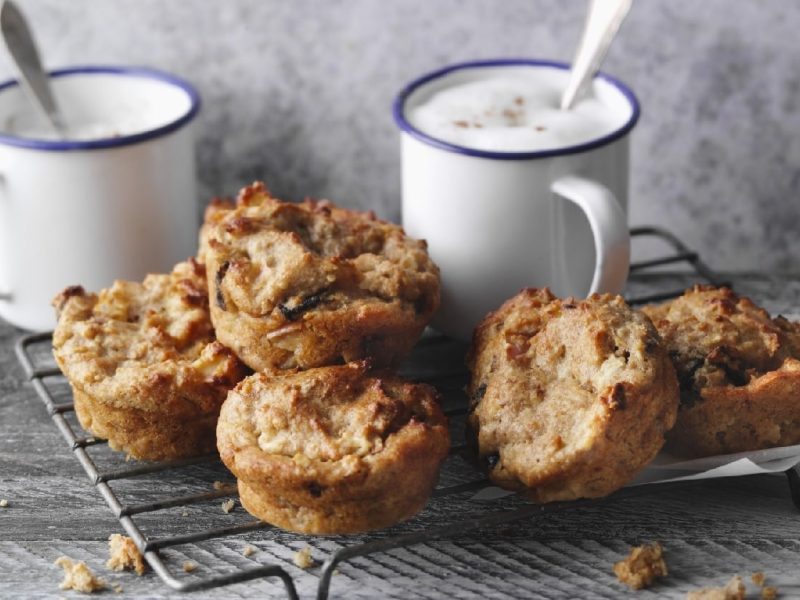 Fünf gebackene Müsli-Muffins auf einem Gitter und zwei helle Kaffeetassen im Hintergrund. Alles auf einem Holztisch und vor grauer Wand.