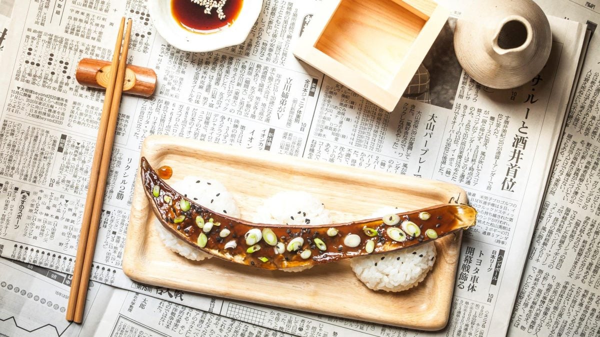 Nasu Dengaku japanische Aubergine mit Miso auf Holzbrett mit Reis und Sesam. Daneben Holzessstäbchen auf Stäbchenhalter. Oben Ponzu-Sauce und Holzschälchen und Sake. Angerichtet auf japanischer Zeitung. Aufnahme aus der Vogelperspektive.