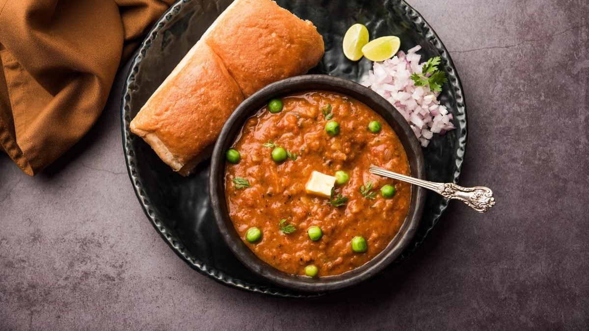 Pav Bhaji, indisches Streetfood, in einer Schale, daneben ein Milchbrötchen, rote Zwiebeln und Limettenspalten. Alles in der Draufsicht.