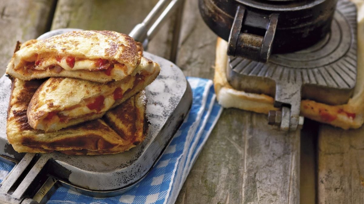 Pizza Jaffles auf einem gusseisernen Waffeleisen. Das Ganze liegt auf einem blauen Küchentuch auf einem hölzernen Untergrund.