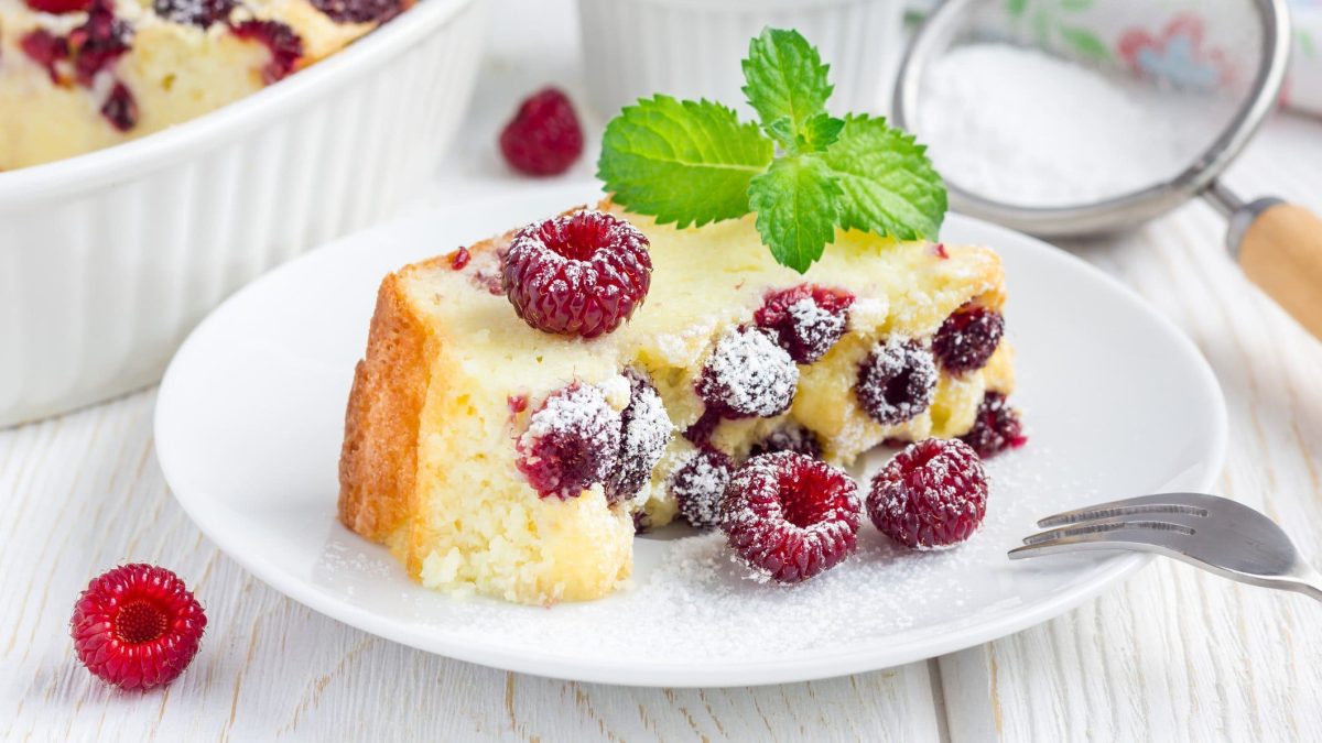 Quark-Vanille-Auflauf mit Himbeeren auf einem weißen Teller. Daneben eine Gabel und ein Sieb mit Puderzucker.