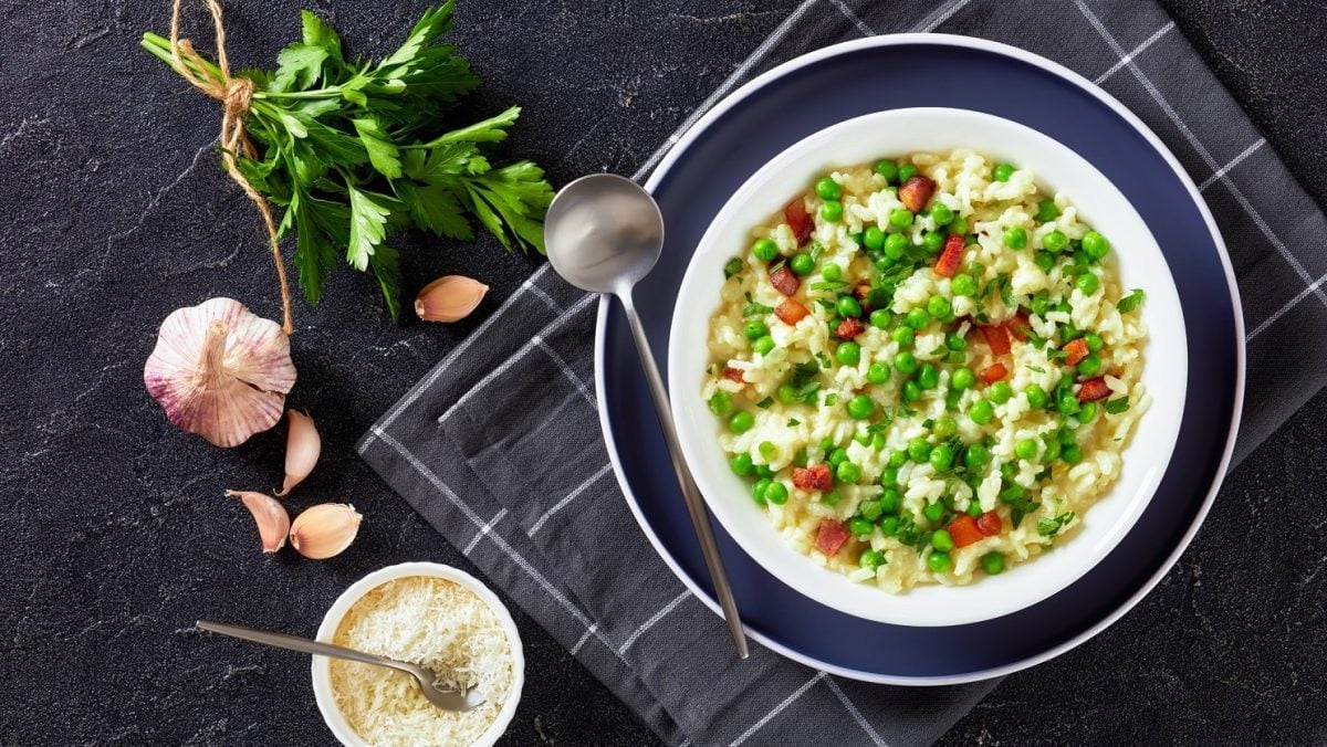 Risi Bisi 80er Retro Rezept in weißem Teller auf dunklem Holz auf grauem Untergrund. Daneben Parmesan in Schüssel, Knoblauch und frische Petersilie. Aufnahme aus der Vogelperspektive