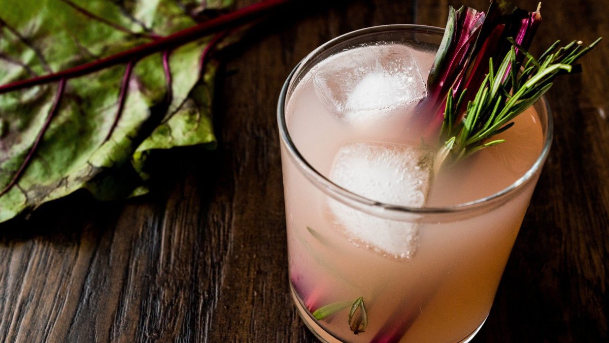 Rhabarber Fizz Cocktail mit Pisco mit Rosmarinzweig und Rhabarber Stange auf dunklem Holz. Daneben ein Blatt Rhabarber. Draufsicht.