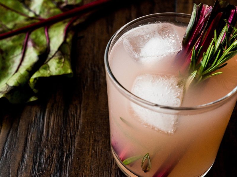 Rhabarber Fizz Cocktail mit Pisco mit Rosmarinzweig und Rhabarber Stange auf dunklem Holz. Daneben ein Blatt Rhabarber. Draufsicht.