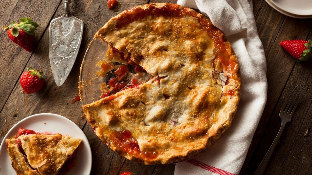 Ein Rhabarber-Pie auf einem Holztisch mit Erdbeeren und einem Tuch. Unten links im Bild noch ein kleiner Teller, mit einem Stück vom Pie.
