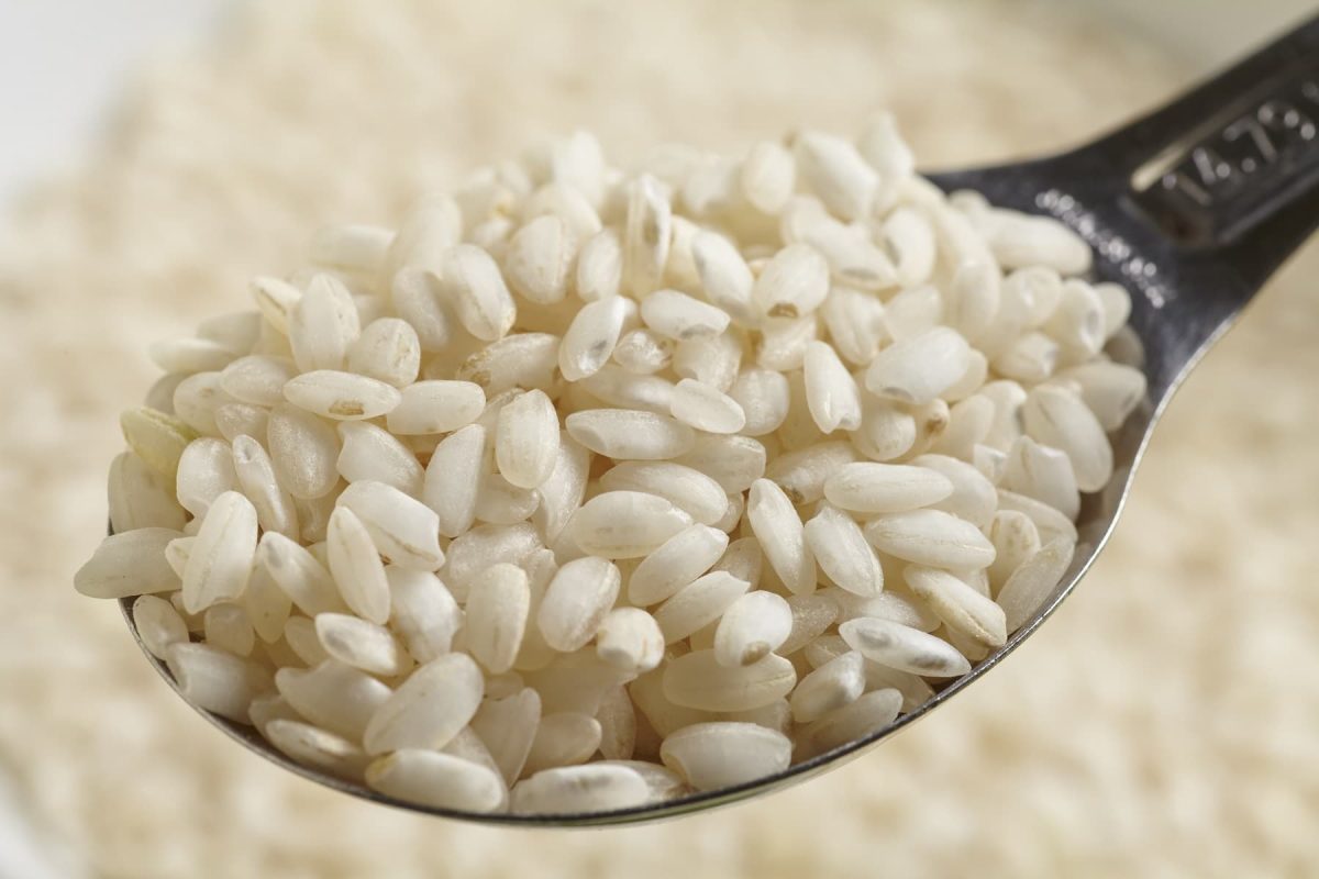 Auf einem Holzlöffel liegt Risotto-Reis. Im Hintergrund ist ebenfalls die Reissorte aus Italien zu sehen.