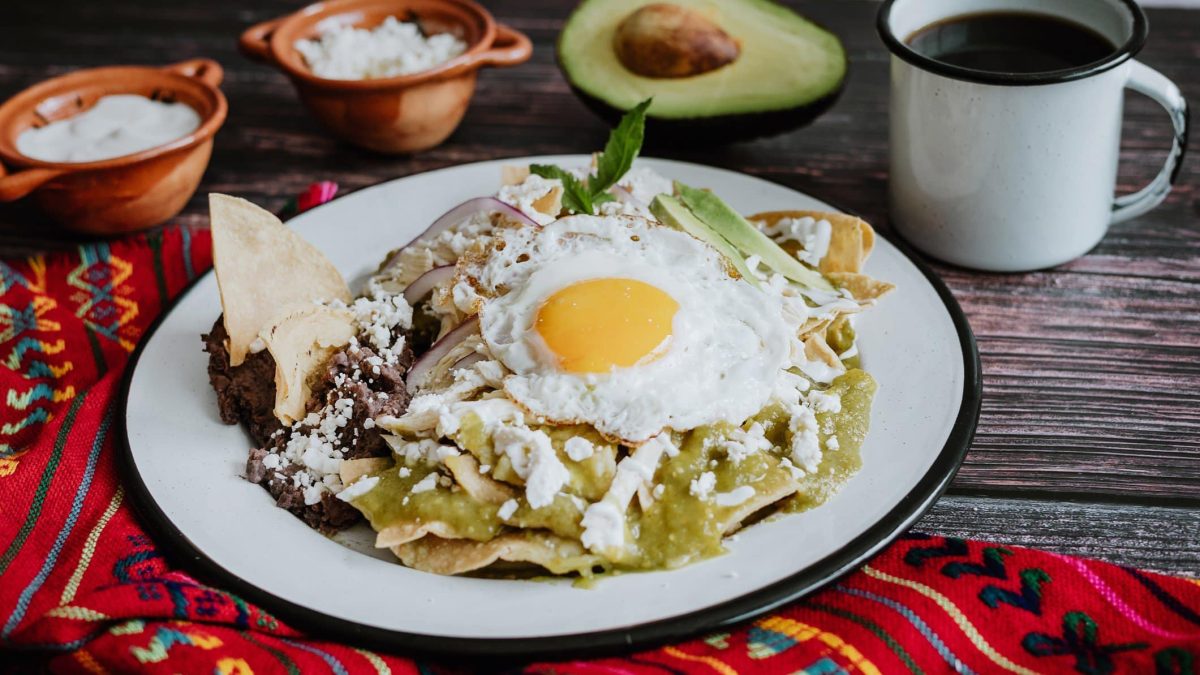 Salsa Verde Baked Eggs auf weißem Teller und grauem Untergrund. Daneben folkloristisches Tuch, Schalen mit geriebenem weißem Käse, eine halbe Avocado und eine Tasse Kaffee.
