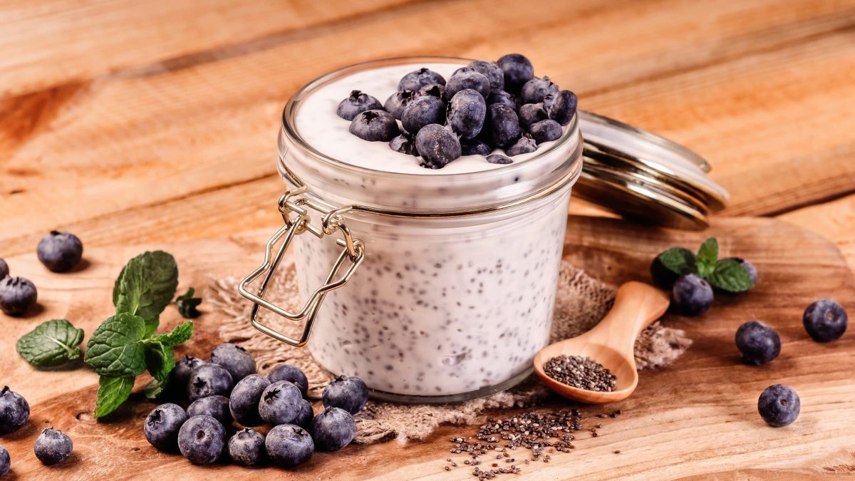 Schnelle Frühstücksbowl mit Blaubeeren und Chiasamen in Einmachglas. Daneben Minze, Blaubeeren und Chiasamen auf Holztisch. Rechts Holzlöffel. Frontalansicht.