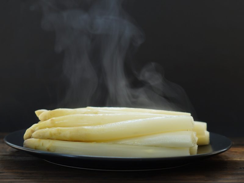 Dampfender weißer Spargel auf einem schwarzen Teller mit dunklem Hintergrund.