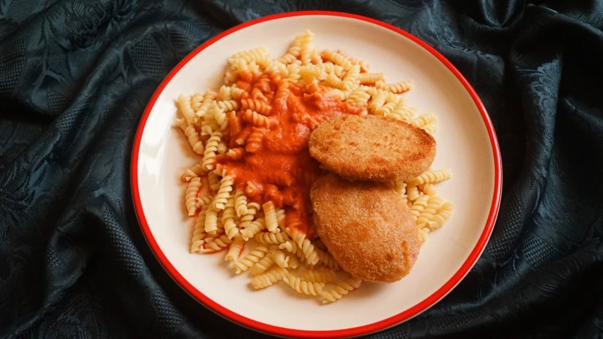 Ein weißer Teller mit rotem Rand, auf dem Spirelli, ein Klecks Ketchupsauce und zwei panierte Jagdwurstscheiben platziert sind. Der Untergrund ist samtig schwarz.