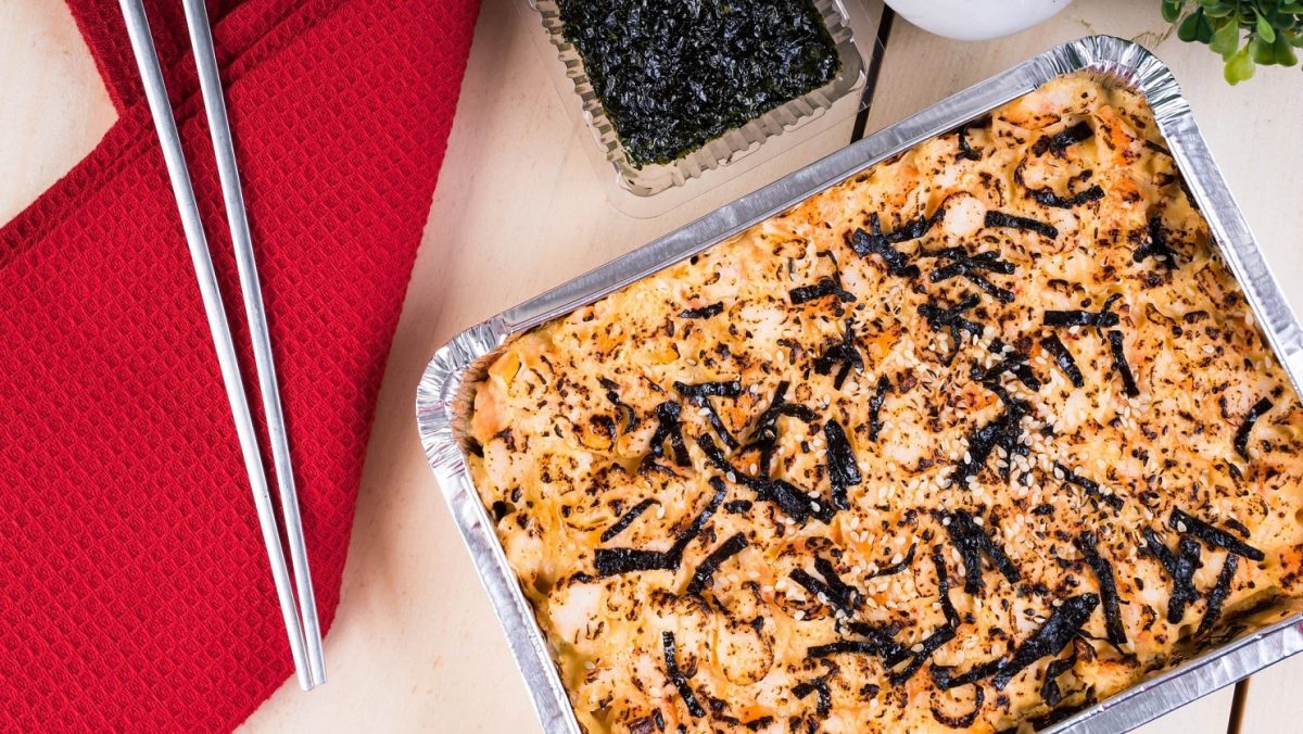 Sushi Bake Sushi aus dem Ofen auf hellem Untergrund. Daneben Metall-Essstäbchen auf rotem Tuch. Oben Schale mit Nori-Streifen. Daneben Saucen. Aufnahme aus der Vogelperspektive.