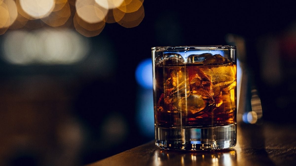 The Godfather Shortdrink aus der Pate auf Holztisch. Im Hintergrund Lichter und Umrisse unscharf zu erkennen. Frontalansicht.