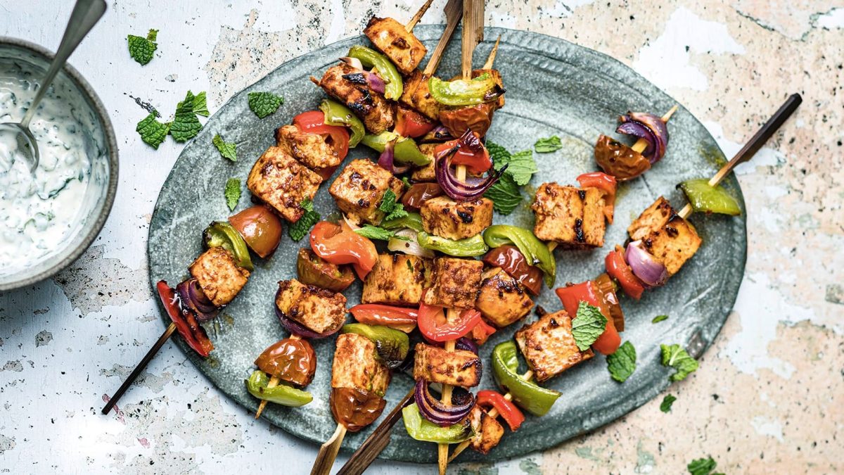 Ein grauer Teller mit Tofu-Kebab-Spießen, die mit Minze bestreut sind, daneben eine Schale mit Joghurtsauce.