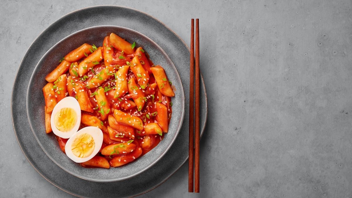 Eine Schüssel mit koreanischem Tteokbokki und einem Paar Stäbchen auf einem grauen Hintergrund.