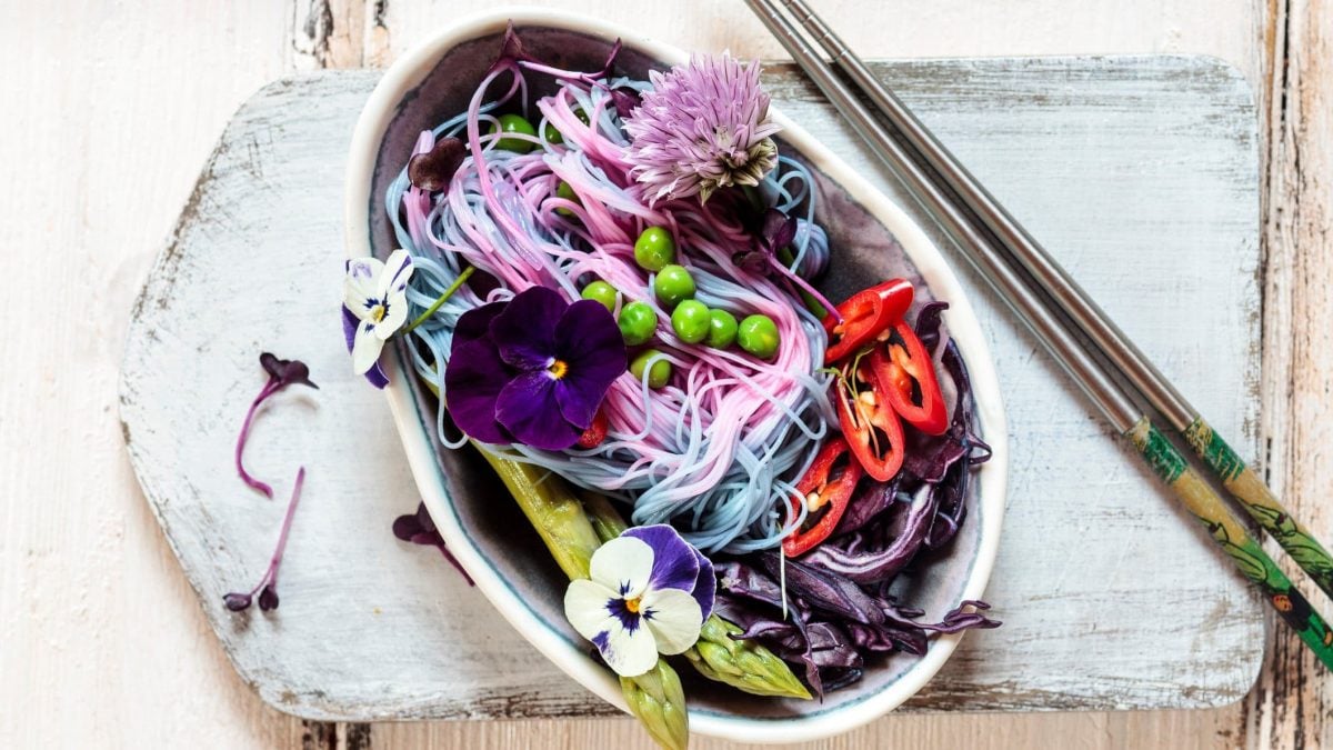 Eine ovale Schale mit den Unicorn Noodles und verschiedenem Gemüse wie Spargel, Rotkohl, Erbsen und dekorativen Blüten.