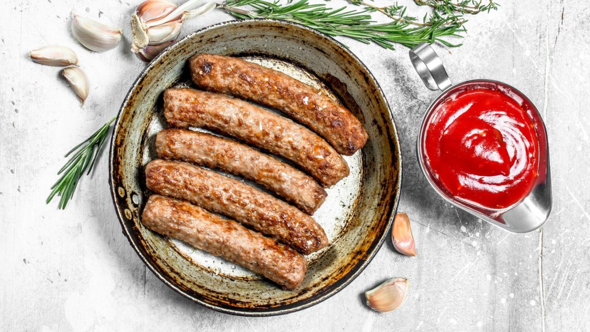 Vegane Bratwürste auf einem Teller, daneben Knoblauch und Kräuter sowie ein Kännchen rote Grillsauce, in der Draufsicht.
