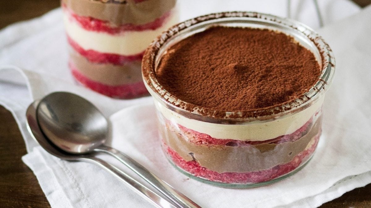 Zwei Gläser des Schichtdesserts auf einer weißen Tischdecke mit zwei silbernen Löffeln im Vordergrund.