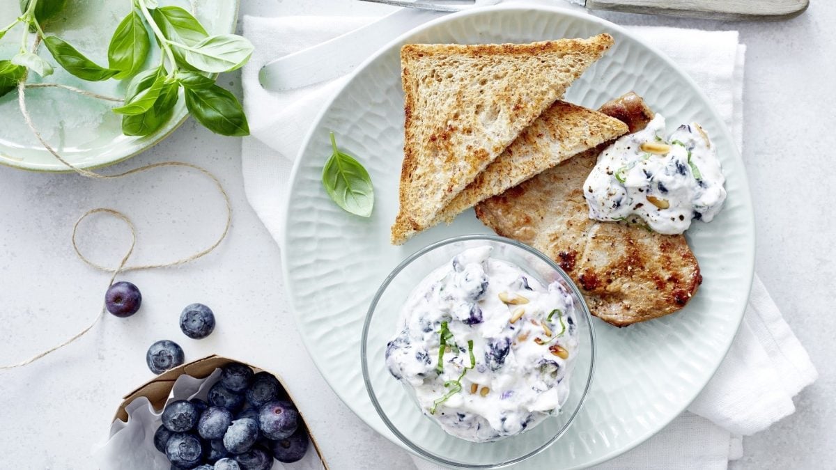 Kalbsschnitzel mit Blaubeer-Dip, bestehend aus Quark, Frischkäse und Pinienkernen, garniert mit frischem Basilikum und gereicht mit getoastetem Toast auf einem hellen Teller. Dazu sind frischen Blaubeeren und Basilikum gereicht.