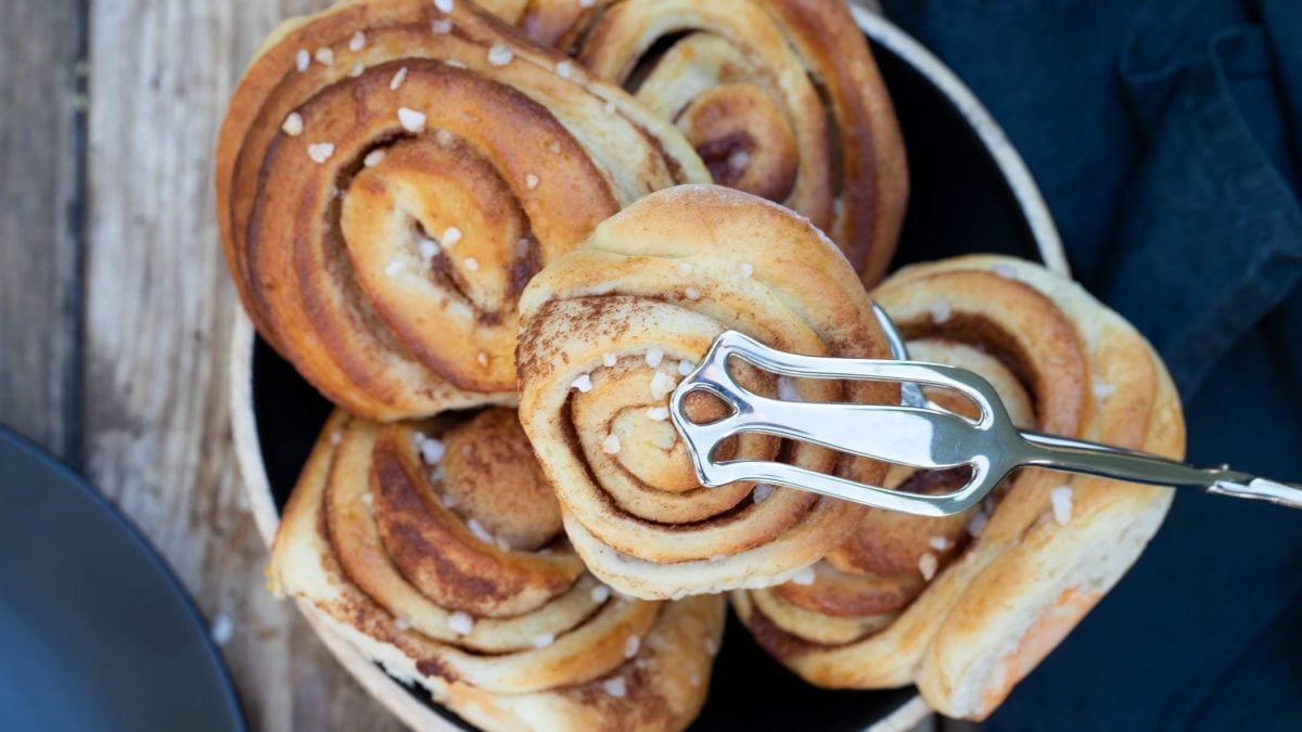 Kanelbullar mit Hagelzucker in einer Schüssel. Eine Zimtschnecke wird mit einer Zange hochgehoben.