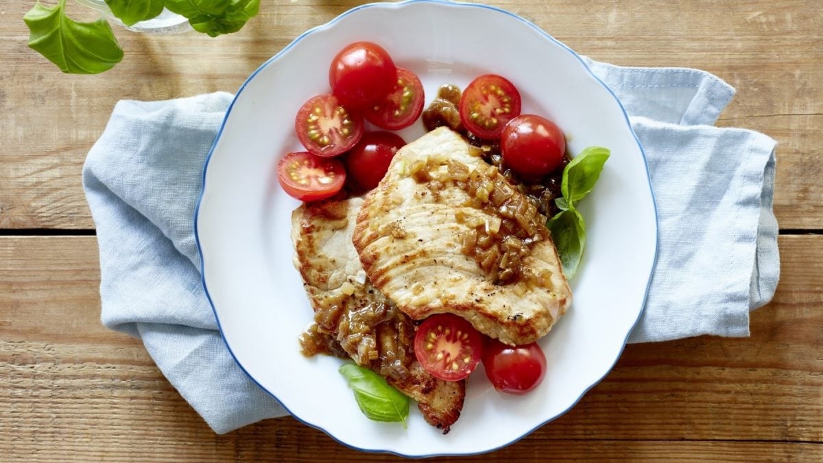 Einfaches Minischnitzel mit Tomate und angerösteten Schalotten, serviert auf einem weißen Teller auf einem Holztisch, garniert mit frischem Basilikum.