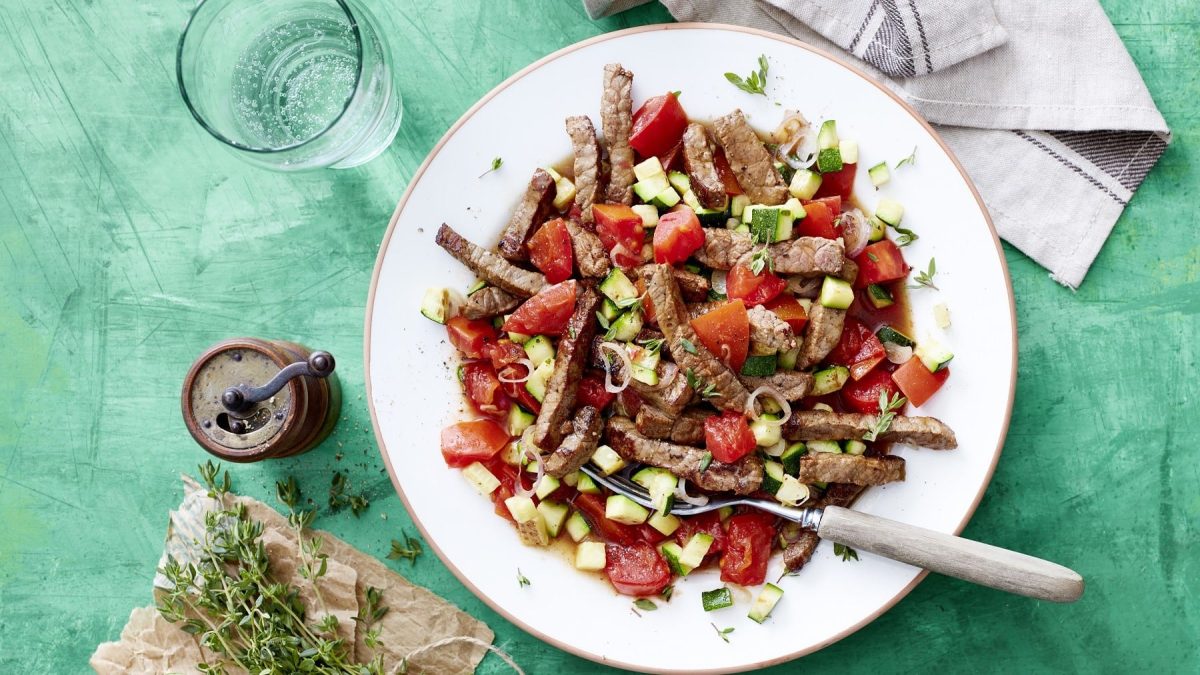 In Streifen geschnittenes Schnitzel mit Tomaten-Zucchini-Sugo und frischem Thymian, serviert in einem weißen Teller auf einem türkisenen Untergrund.