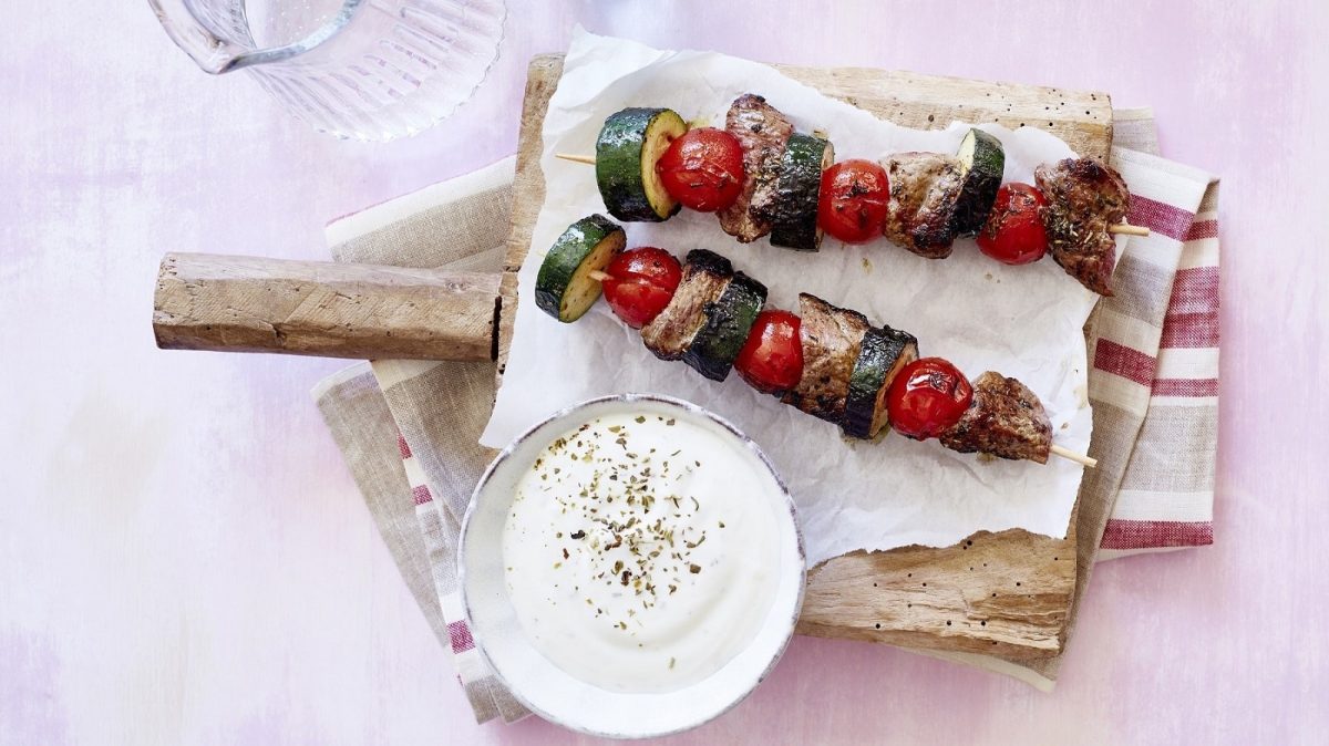Auf einem Holzbrett liegen Steak-Spieße mit Zucchini und Kirschtomaten. Daneben steht ein Dip.