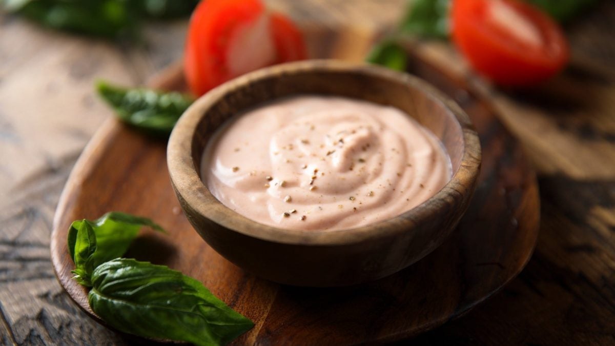 Thousand-Island-Dressing in dunkler Schale auf Holzbrett. Daneben liegen Basilikum und Tomaten.