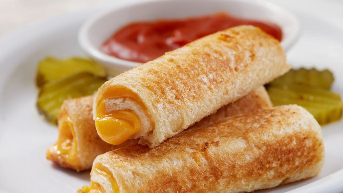 Mehrere gerollte Toast-Brote mit Käsefüllung auf weißem Teller. Daneben ein Tomaten-Dip und Gewürzgurken.