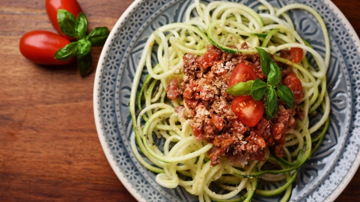 Auf einem Teller liegt eine Portion Zoodles mit Tofu-Bolognese. Sie ist mit vegetarischem Hack und Tomaten garniert.