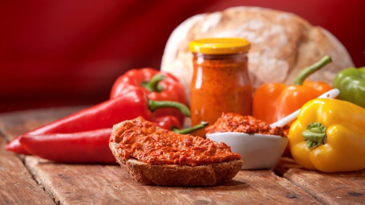 Ajvar auf einer Scheibe Brot vor Gemüse und einem Glas mit Ajvar. Im Hintergrund Brotlaib. Frontalansicht.