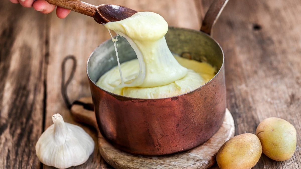 Ein kupferner Topf auf einem Holzbrett mit Knoblauch und Kartoffeln an der Seite. Eine Hand zieht mit einem Kochlöffel etwas von dem Aligot in die Länge.