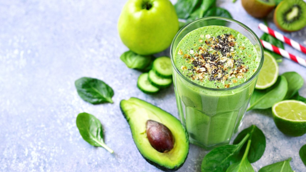 Ein grünes Glas mit dem Smoothie auf grauem Untergrund. Drum herum viel grünes Obst und Gemüse.