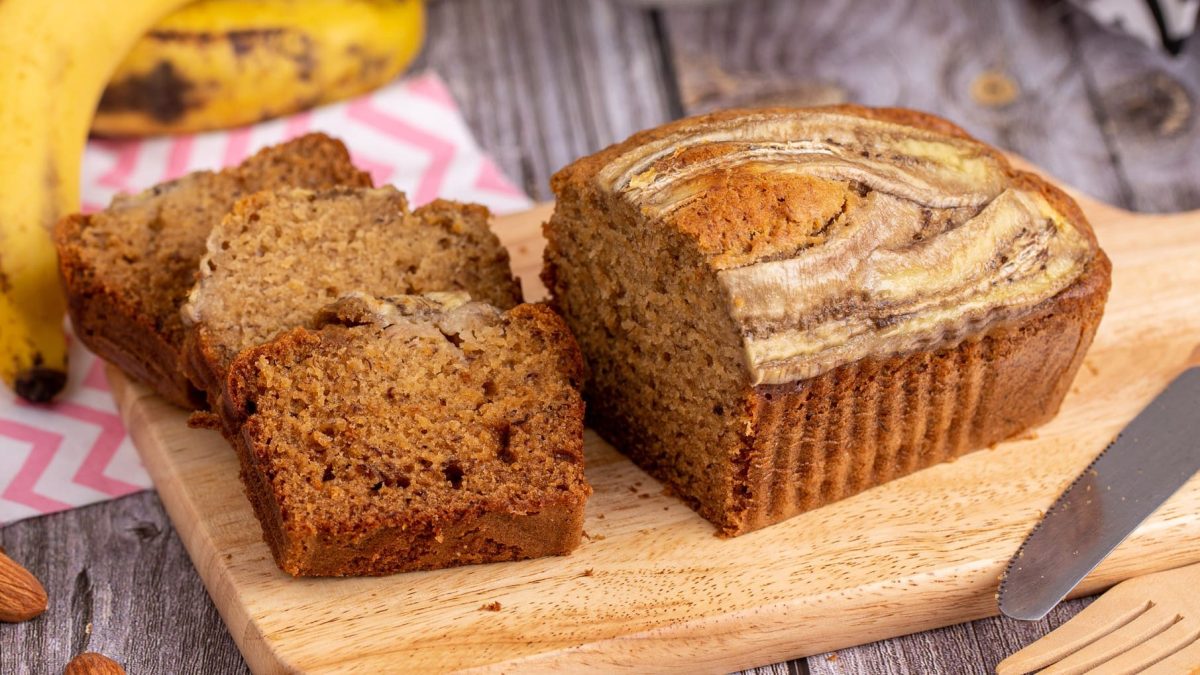 Selbst gemachtes Bananenbrot auf Holzbrett und grauem Holztisch. Oben reife Bananen und rot-weißes Stofftuch. Unten rechts Messer. Drei Scheiben geschnitten auf dem Brett trappiert. Frontalansicht.