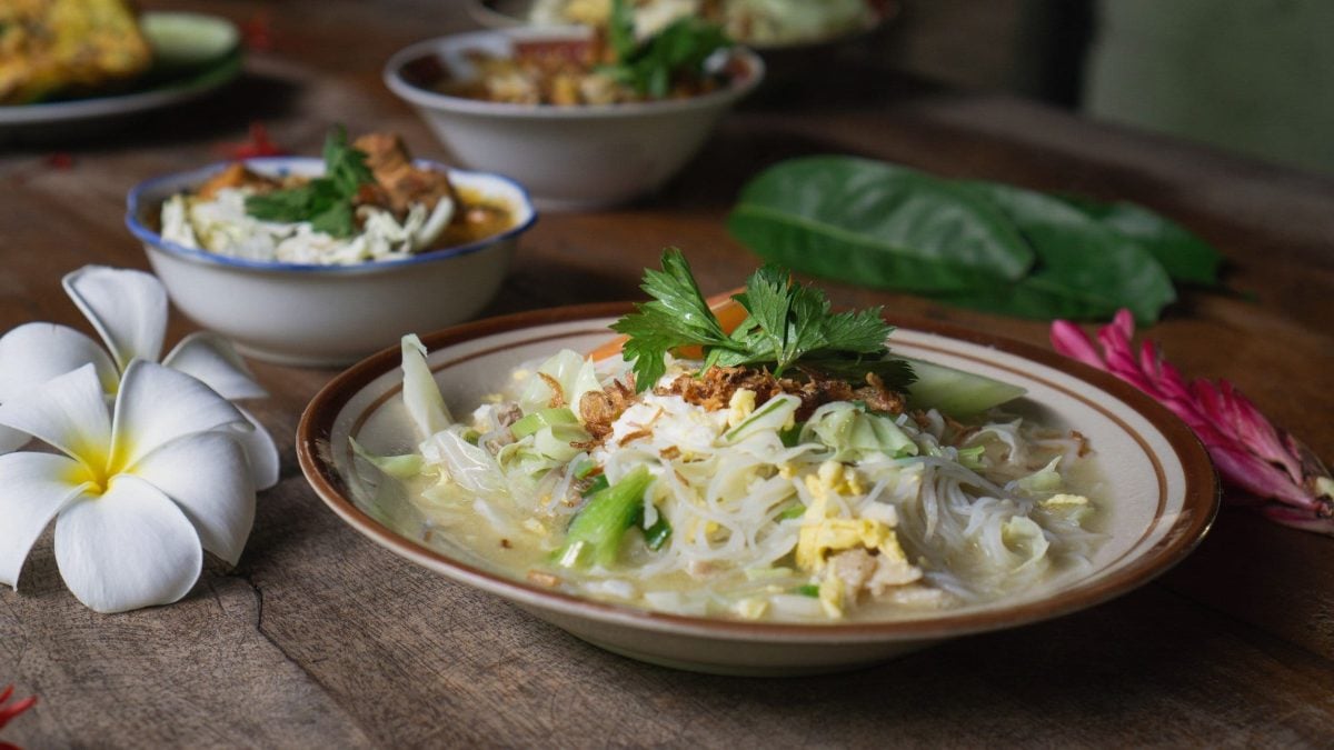 Bihunsuppe Bihun Sup Ayam in Suppenteller auf Holztisch. Blüten und Schalen mit Deko im Hintergrund.