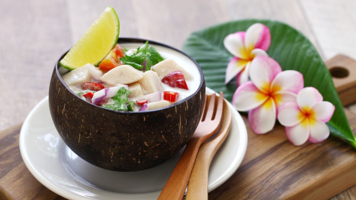 Thuna-Ceviche mit Kokosmilch und Chili in Kokosschale auf weißem Teller auf Holzbrett. Daneben Holzbesteck und Blütendeko. Draufsicht.