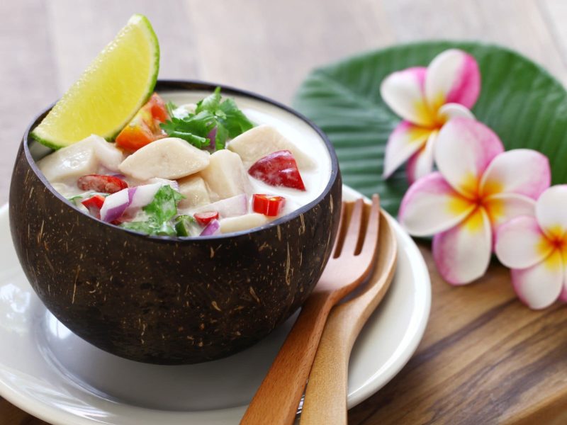 Thuna-Ceviche mit Kokosmilch und Chili in Kokosschale auf weißem Teller auf Holzbrett. Daneben Holzbesteck und Blütendeko. Draufsicht.
