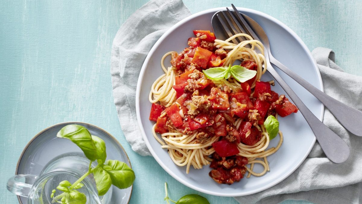 Chili-Pasta mit Hack und Paprika auf einem weißen Teller. Daneben Besteck und Basilikum.