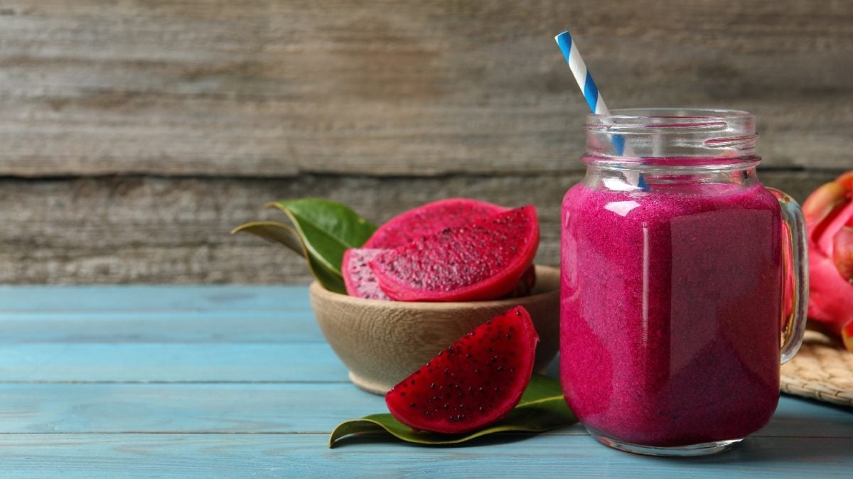 Ein Glas mit dem Drachenfrucht Smoothie auf einem blauen Holztisch mit angeschnittener Drachenfrucht im Hintergrund.