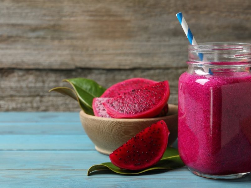 Ein Glas mit dem Drachenfrucht Smoothie auf einem blauen Holztisch mit angeschnittener Drachenfrucht im Hintergrund.
