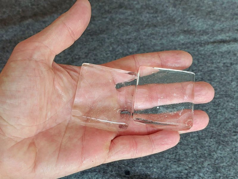 Eine Hand mit zwei durchsichtigen Eiswürfeln auf grauem Untergrund.