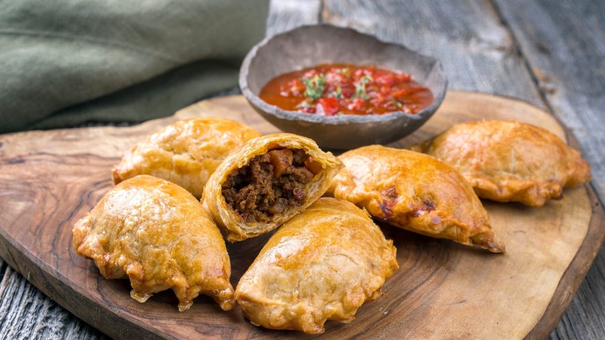 Fünf Empanadas auf einem Holzbrett und grauem Hintergrund. Hinter dem Brett ist eine Dip-Schale mit einer roten Salsa.